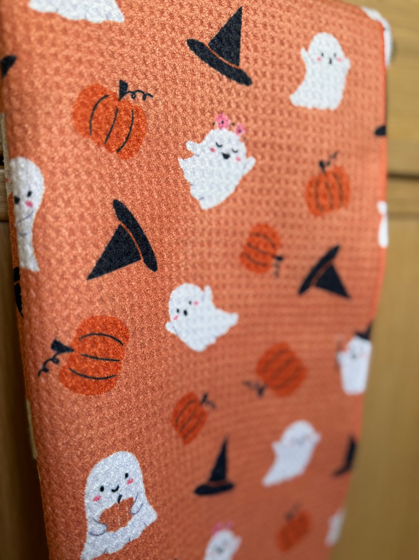 towel with Halloween-themed pattern featuring ghosts, pumpkins, and witch hats on an orange background.