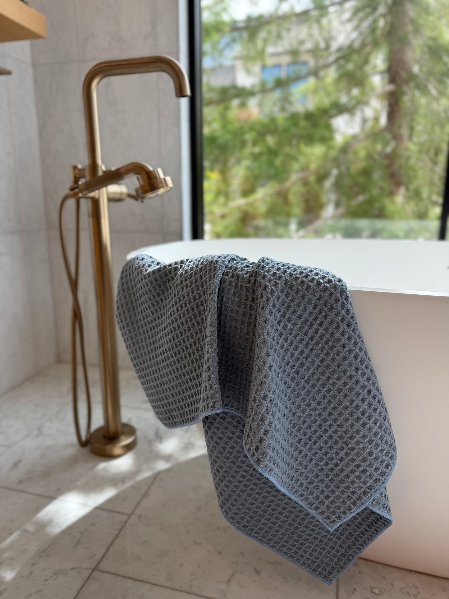 light blue waffle-textured towel draped over a white bathtub in a bathroom with gold fixtures and a window view.