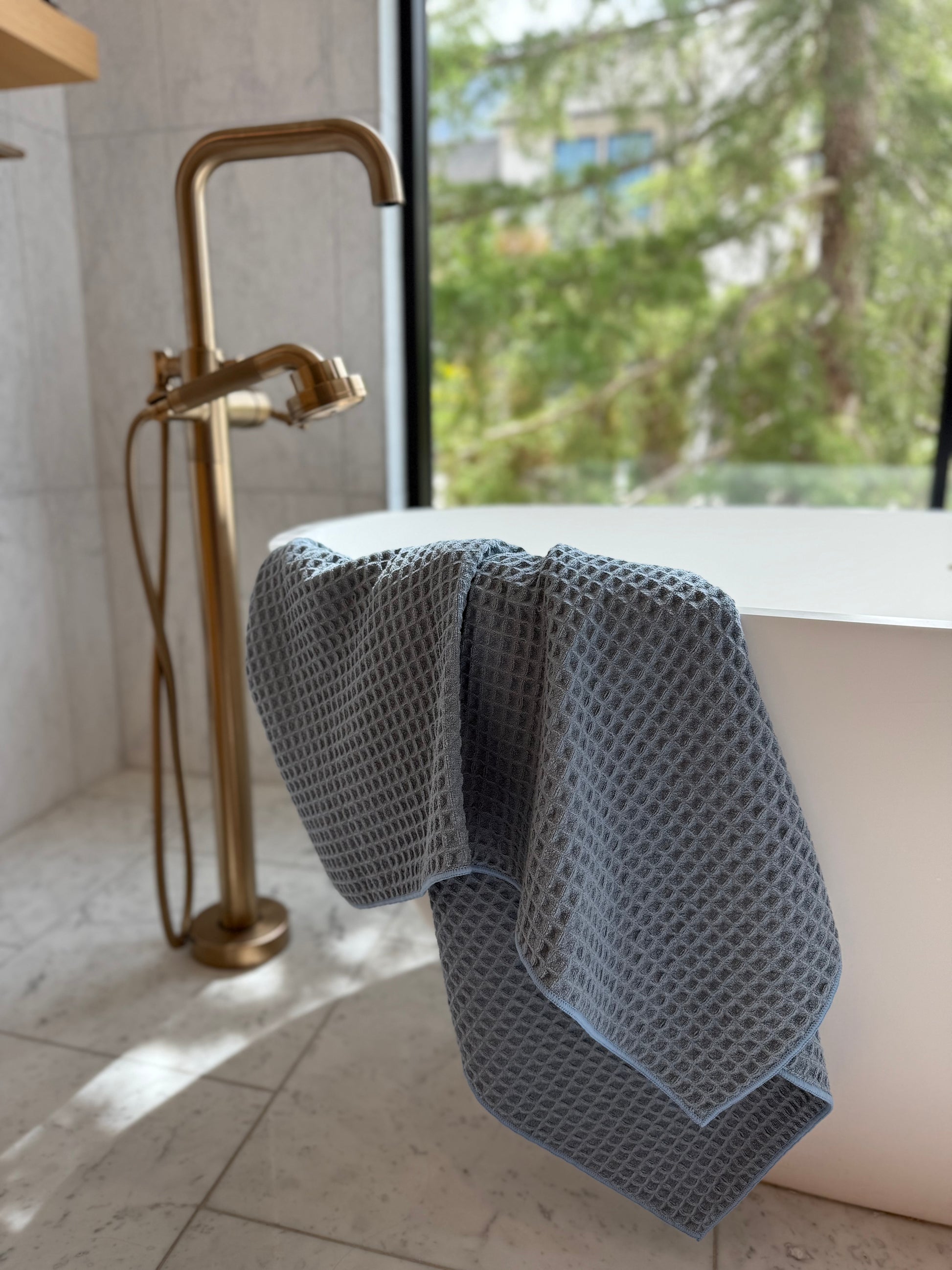 light blue waffle-textured towel draped over a white bathtub in a bathroom with gold fixtures and a window view.